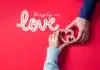 Two hands hold a wooden heart together against a vibrant red background with the words "deeply in love with you" written in cursive, symbolizing a shared affection and unity of hearts.