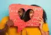 A cheerful couple playfully peeking through a heart-shaped red sign adorned with the word "love" in various sizes and fonts, against a vivid blue background, symbolizing the 5 Love Languages and