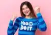 A young woman with a joyful expression, wearing a blue and white striped shirt with the motivational phrase "you can do it" printed on it, makes an enthusiastic fist pump gesture against a soft pink background