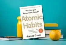 A still life composition featuring the bestselling self-help book 'Atomic Habits' by James Clear, alongside a bright yellow cleaning spray bottle, a stack of colorful books, and a fresh orange, all set