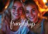 Two smiling young women with blonde hair sit close together, illuminated by warm lighting. The word "strong friendship" is elegantly written across the image, highlighting their bond. The background is softly blurred, focusing on their happy expressions.