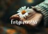 A close-up of an open hand holding a white daisy with a yellow center. The background is blurred with warm tones. The word "Forgiveness" is elegantly scripted in white across the image, complementing the serenity. The person appears to be wearing a textured sweater.