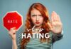 A woman with red hair and a striped shirt holds up a red stop sign with the word "HATE" on it. With a serious expression, she extends her other hand forward, palm facing out. The text "How to Stop Hating Someone Who Hurt You" is overlaid on the image, emphasizing her message against hate.