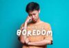 A person wearing a tan shirt stands in front of a teal background, looking down with a thoughtful expression while resting their chin on their right hand. The text "How-to-Embrace-Boredom" is overlaid on the image in white lettering.