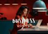 A woman in a red blazer, seemingly addicted-to-busyness, works intently on a laptop in a modern, dimly-lit space with hanging lamps. Text overlay reads "BUSYNESS signs.