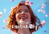 A young woman with short red hair is smiling and looking up. She is surrounded by falling pink flower petals against a bright blue sky. The text "how-to-become-mature-emotionally" is overlaid on the image in white, playful cursive font.